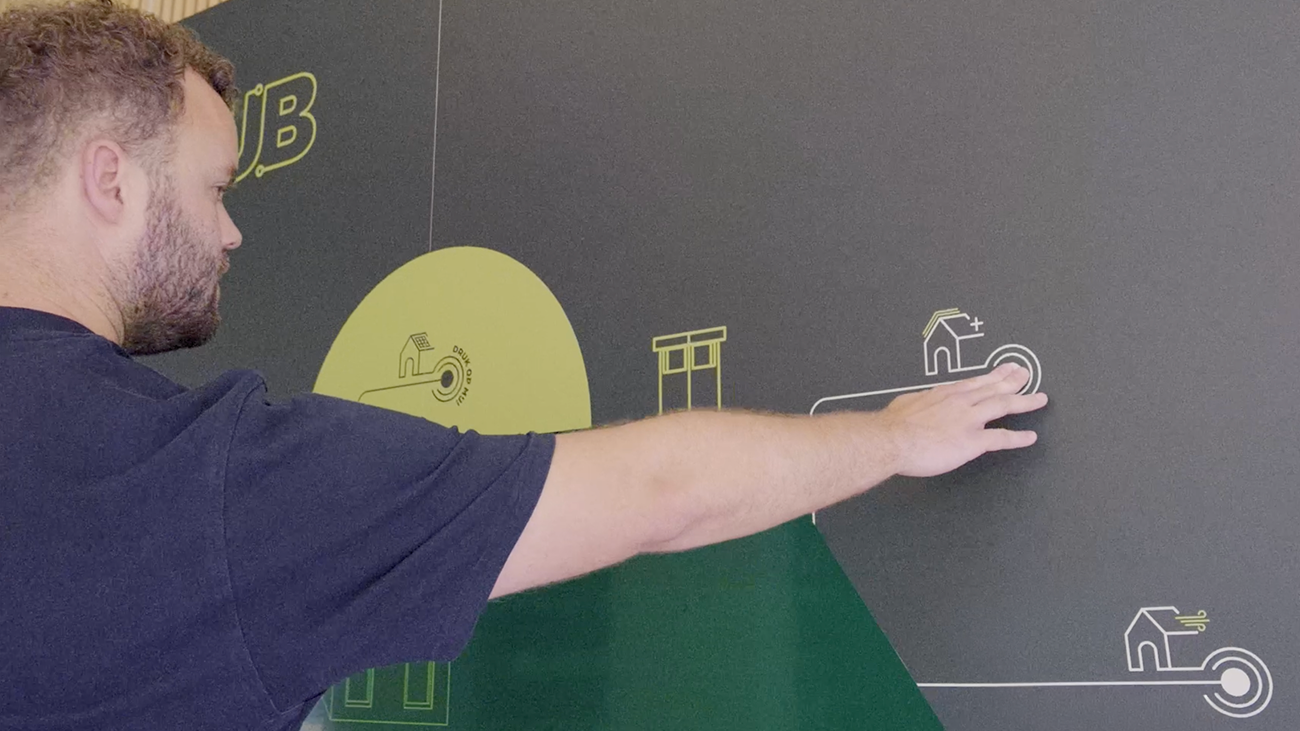Close-up van een man die met uitgestrekte arm een icoon op een interactieve wand aanraakt, onderdeel van een visuele uitleg over duurzame woningoplossingen.
