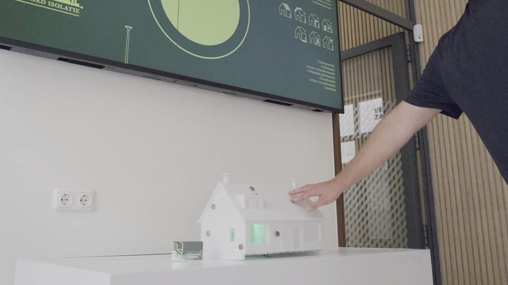 Hand van een man die een wit schaalmodel van een huis aanraakt op een tafel, met op de achtergrond een digitaal scherm over woningisolatie en verduurzaming.