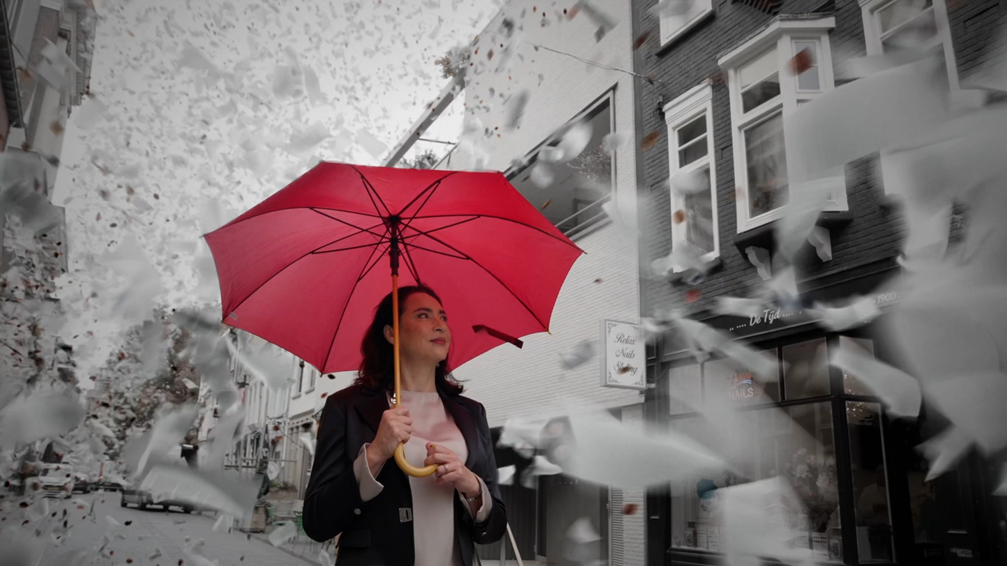 Woman holding a bright red umbrella in a black and white street scene while papers fly around her in the wind.