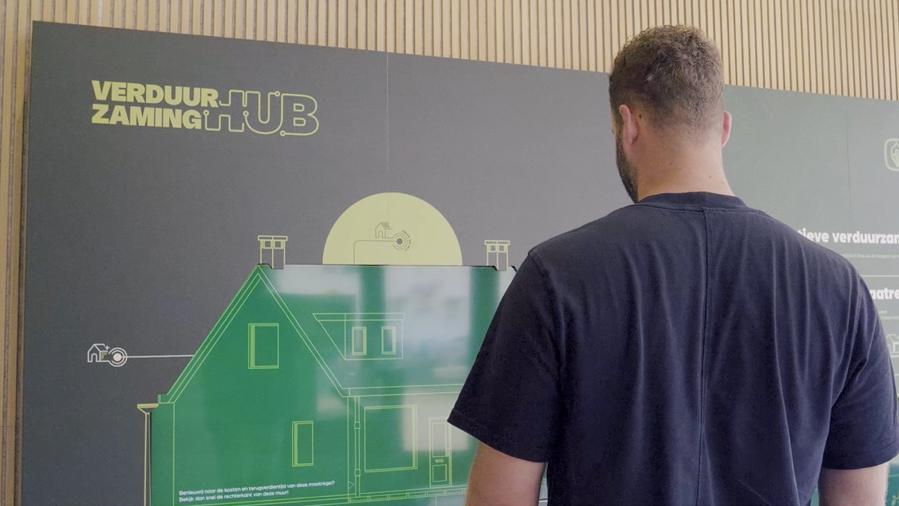 Man staat met zijn rug naar de camera voor een groot interactief scherm in de Verduurzaming Hub, waarop een woning en verduurzamingsopties visueel worden weergegeven.