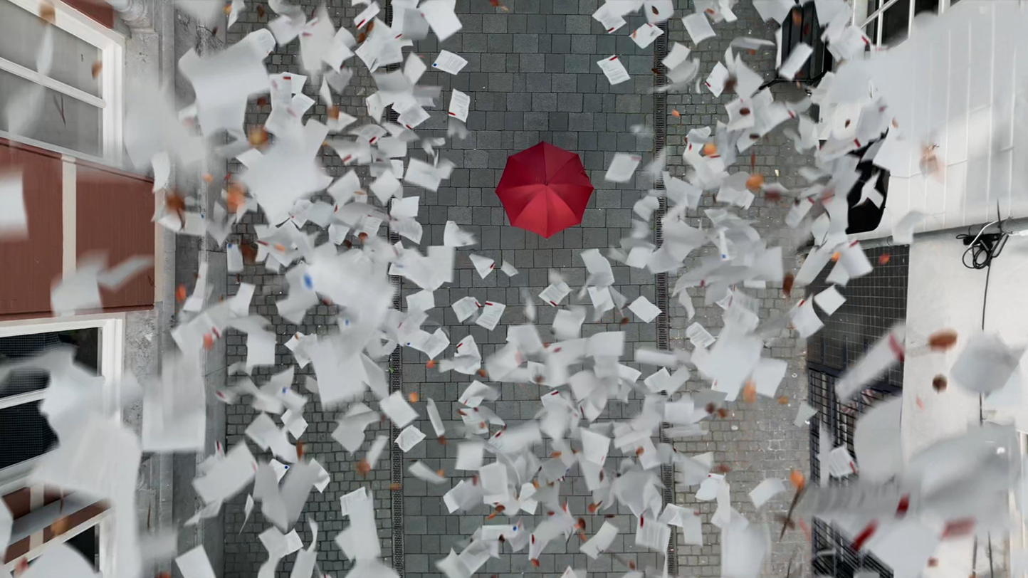 Top-down view of a red umbrella centred in a narrow cobbled street while dozens of white papers blow around in the wind.